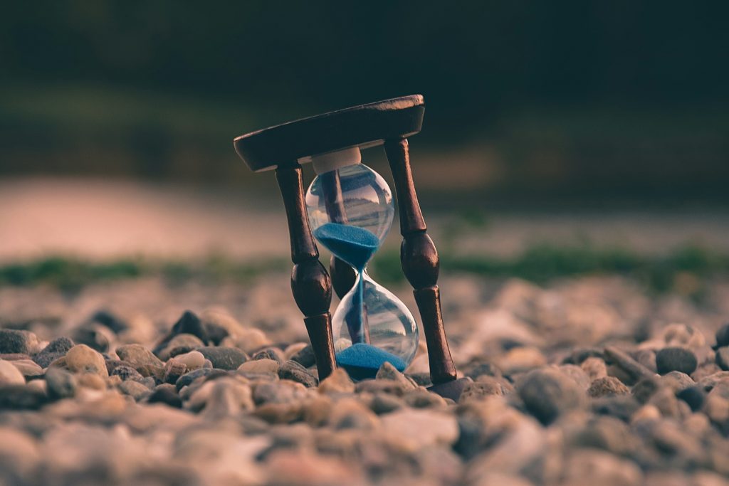 A vintage wooden hourglass with blue sand placed on a rocky surface, symbolising the passage of time and urgency.